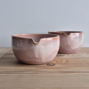 May include: Two ceramic bowls with a light pink and white glaze. Each bowl has a small spout and sits on a wooden surface. The bowls are round with a smooth, glossy finish, and the glaze has a subtle, marbled effect.