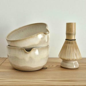 Puede incluir: Dos cuencos de cerámica blanquecina apilados, cada uno con un pico curvo. Un batidor de matcha de bambú se encuentra junto a los cuencos. Los cuencos y el batidor están sobre una superficie de madera clara, con un fondo blanco liso.