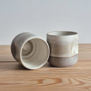 May include: Two ceramic cups with a speckled off-white and earthy brown finish. One cup is positioned on its side, revealing the interior, while the other stands upright. The exterior features a textured, matte surface with a gradient color transition.