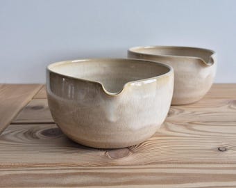 May include: Two beige ceramic bowls with a pouring spout, set on a light-colored wooden surface. The bowls have a glossy finish and a rounded shape. The background is a plain, light-colored wall. The bowls are likely for kitchen use.