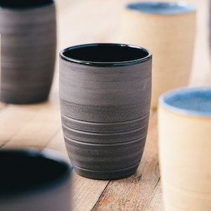 Könnte beinhalten: Eine Sammlung handgefertigter Keramikbecher. Der vordere Becher ist dunkelgrau mit horizontalen, geätzten Linien und einem schwarzen Rand. Weitere Becher sind sichtbar, einige in cremefarben und mit blauen Rändern. Der Hintergrund ist eine helle Holzoberfläche.