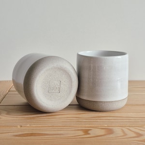 Op de afbeelding: Twee keramische mokken met een wit geglazuurd bovendeel en een ongeglazuurde, gespikkelde beige basis. De ene mok staat rechtop, terwijl de andere op zijn kant ligt, wat een vierkante stempel op de bodem met de tekst MUG MUG onthult.