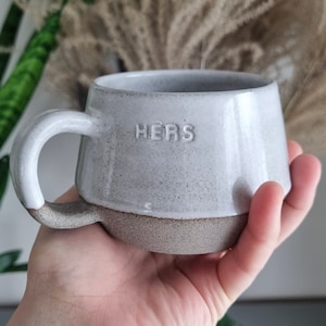 May include: A white ceramic mug with a speckled glaze and a brown rim. The mug has a handle and the word "HERS" embossed on the front.