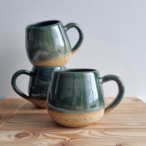 Puede incluir: Tres tazas de cerámica hechas a mano apiladas sobre una superficie de madera. Las tazas son de color verde oscuro con una base moteada de color canela y un asa curva. Las tazas tienen un acabado brillante y una forma redondeada.