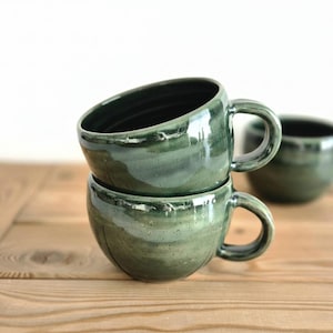 Puede incluir: Dos tazas de cerámica hechas a mano apiladas, con un esmalte verde brillante y moteado. Cada taza tiene un cuerpo redondeado, un asa curva y un interior oscuro. Una tercera taza es visible en el fondo. Las tazas están sobre una superficie de madera.