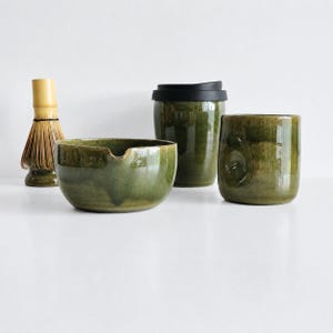 May include: A set of matcha tea accessories including a bamboo whisk, a green ceramic bowl with a pouring spout, a green ceramic lidded tumbler with a black lid, and a green ceramic mug with a textured side indentation for a secure grip.