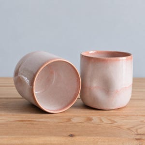 May include: Two ceramic tumblers with a soft, pale pink glaze and a subtle ombre effect that deepens towards the rims. The cups feature a slightly irregular, organic shape with smooth, rounded walls and a wide, open top design.