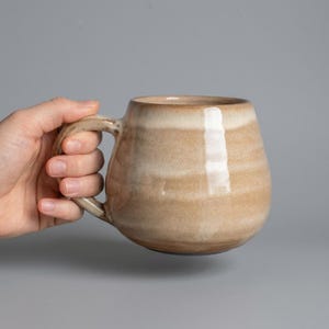 Può includere: Una tazza in ceramica arrotondata con una smaltatura liscia e lucida caratterizzata da fasce orizzontali nei colori beige, marrone chiaro e crema. La tazza presenta un corpo ampio e curvo con un manico robusto integrato sul lato.