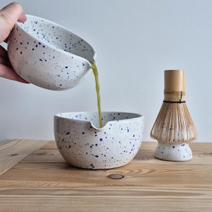 Może przedstawiać: Białe ceramiczne miski do matchy z niebieskimi plamkami. Jedna miska wlewa zielony płyn do drugiej miski. Bambusowa miotełka do matchy stoi w pobliżu na drewnianej powierzchni. Miski mają dzióbek.