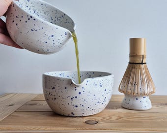 May include: White ceramic matcha bowls with blue speckles. One bowl is pouring green liquid into another bowl. A bamboo matcha whisk sits nearby on a wooden surface. The bowls have a spout for easy pouring.