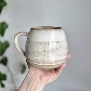 Könnte beinhalten: Eine handgefertigte Keramiktasse mit runder Form und einem stabilen Henkel. Die Tasse hat eine gesprenkelte, cremefarbene Glasur mit Bändern in Beige und Braun. Der Boden ist aus naturbelassener, unglasierter Keramik.