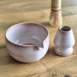 May include: A light pink ceramic matcha bowl with a spout, a matching chawan stand, and a bamboo whisk. The bowl and stand have a gradient colour effect. The items are on a wooden surface.