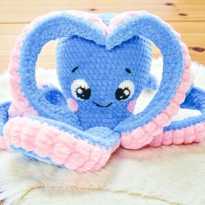 Peut inclure: Une peluche pieuvre au crochet bleue et rose faite à la main. La pieuvre a de grands yeux expressifs, des joues roses et une bouche souriante. Les tentacules sont bleues avec des accents roses, et certaines forment un cœur. Le jouet est sur une surface blanche et moelleuse.