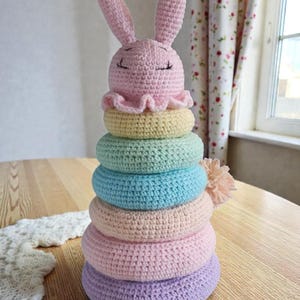 Op de afbeelding: Een handgemaakt gehaakt stapelspeelgoed met een konijnenkop en gekleurde ringen. Het konijn is lichtroze met gesloten ogen, en de ringen zijn pastelkleuren geel, groen, blauw, roze en paars. Een pompon is bevestigd. Het speelgoed staat op een houten tafel.