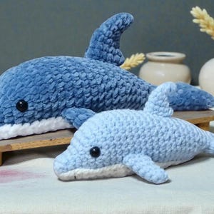 Peut inclure: Deux dauphins en peluche crochetés, dans des tons de bleu. Le plus grand, bleu foncé, repose sur un petit support en bois. Le plus petit est bleu clair. Les deux ont des yeux noirs et un ventre blanc.