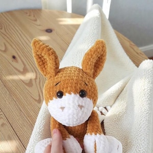 May include: A handmade, plush crochet bunny in shades of brown and white. The bunny has large, upright ears, a white face, and white paws. It is sitting on a soft, white blanket.