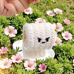 May include: A white crocheted toilet roll cover with an angry face, set amongst green foliage and pink flowers. The handmade cover adds a whimsical touch to bathroom decor.