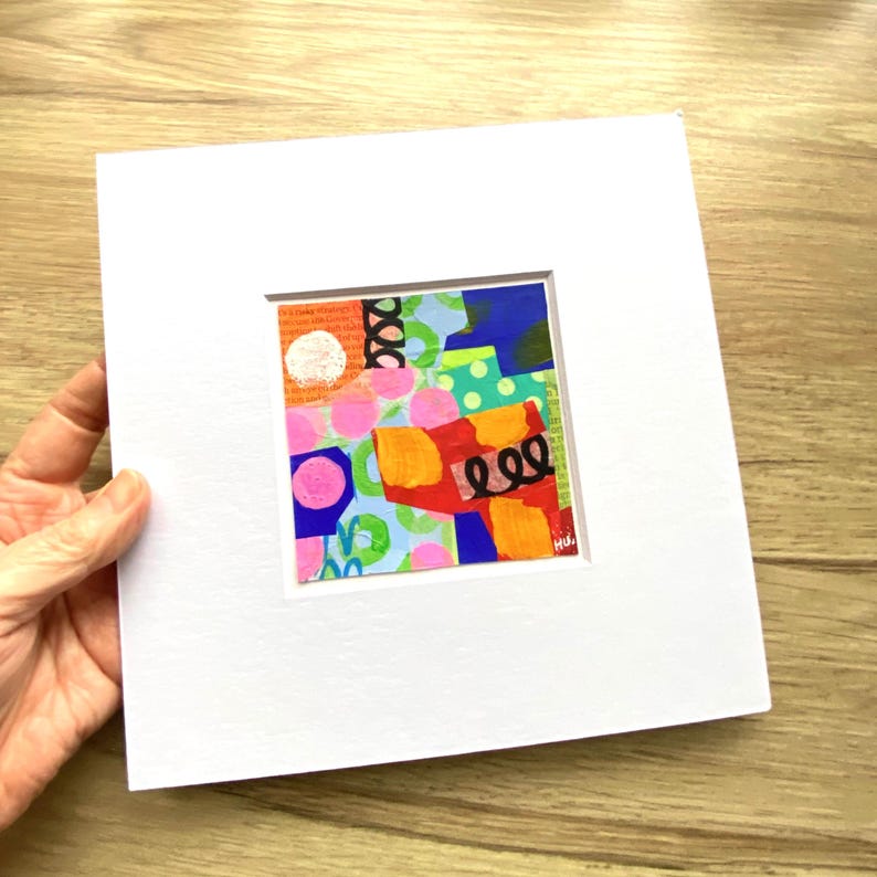 May include: A small, square abstract painting with a white mat. The artwork features a collage of colorful shapes, including pink circles, orange rectangles, and blue and green patches. Black squiggles and a white circle add detail. The artist's signature is in the lower right corner.