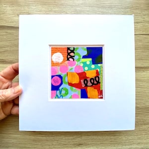 May include: Abstract art piece with a colorful collage of shapes and patterns. The artwork features pink circles, orange rectangles, and blue and green shapes, all set against a white background. The piece is mounted in a white mat.