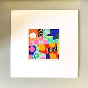May include: Abstract mixed media artwork with a white mat. The artwork features a collage of colorful shapes, including pink circles, blue and yellow blocks, and black squiggles. The background includes text fragments.