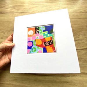 May include: A colorful abstract art piece with a square, matted design. The artwork features a collage of shapes and patterns in various colors, including pink, orange, blue, and green. The piece is set against a white mat.