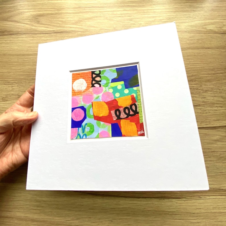 May include: A colorful abstract art piece with a square, matted design. The artwork features a collage of shapes and patterns in various colors, including pink, orange, blue, and green. The piece is set against a white mat.