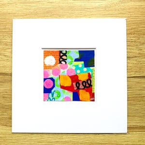 May include: Abstract mixed media artwork with a collage of colorful shapes and patterns. The artwork features a white circle, pink polka dots, and a black swirl on a background of text and various colors. The artwork is matted in a white frame.
