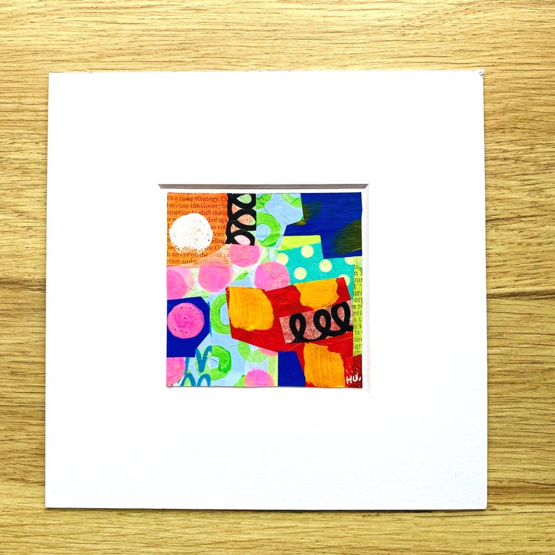 May include: Abstract mixed media artwork with a collage of colorful shapes and patterns. The artwork features a white circle, pink polka dots, and a black swirl on a background of text and various colors. The artwork is matted in a white frame.