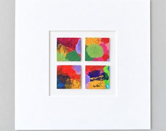 May include: A square white mat frame containing four small square panels arranged in a grid. Each panel features abstract painted designs in vibrant colors including red, yellow, green, blue, and purple with visible brushstroke textures.