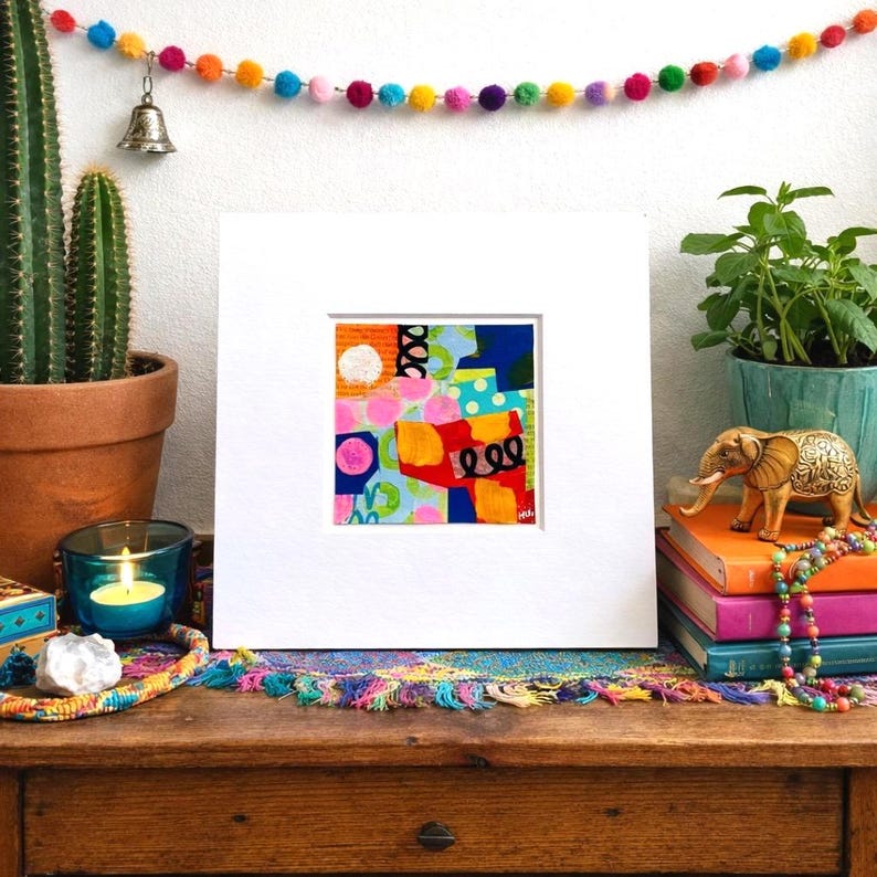 May include: A framed abstract art piece with a colorful design of pink circles, orange shapes, and blue and green backgrounds. The artwork is displayed on a wooden table with decorative items, including a cactus, candle, and a string of colorful pom-poms.