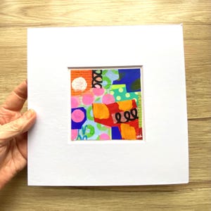 May include: Abstract art piece with a white mat. The artwork features a collage of colorful shapes, including pink circles, orange brushstrokes, and blue and green geometric forms. The piece is signed in the lower right corner.