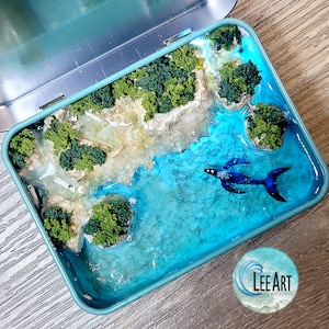 May include: A miniature ocean diorama in a teal tin. The scene includes a blue whale in turquoise water, surrounded by miniature islands with green trees and sandy beaches. The diorama is a unique art piece, a creative craft.