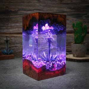 Puede incluir: Un bloque de resina transparente con una base de madera, que contiene un remolino de resina p&uacute;rpura y azul con un &aacute;rbol de metal plateado en su interior. El bloque est&aacute; iluminado desde el interior, proyectando un brillo p&uacute;rpura.