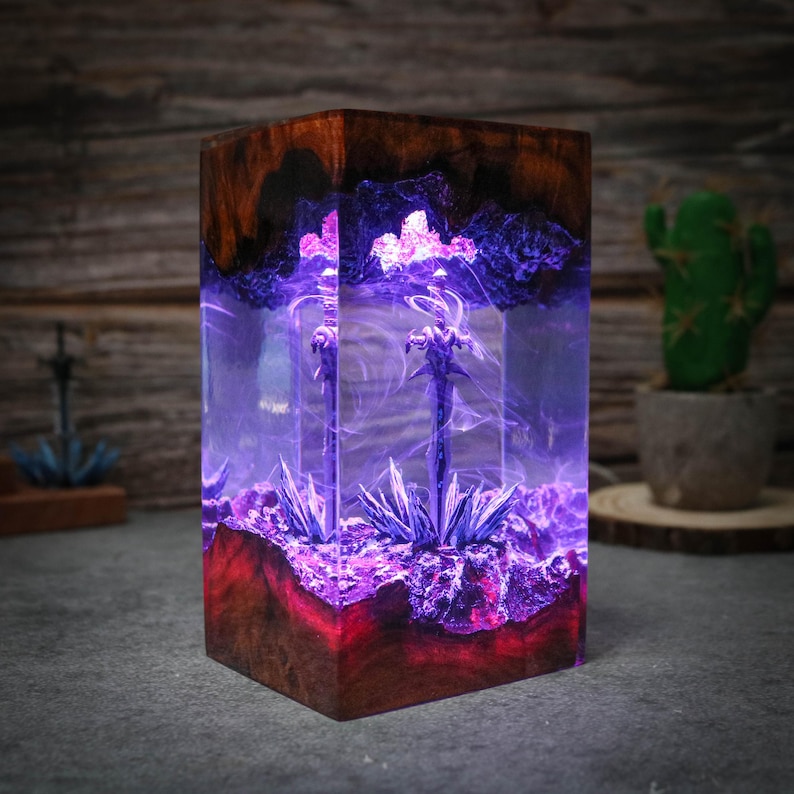 Puede incluir: Un bloque de resina transparente con una base de madera, que contiene un remolino de resina p&uacute;rpura y azul con un &aacute;rbol de metal plateado en su interior. El bloque est&aacute; iluminado desde el interior, proyectando un brillo p&uacute;rpura.