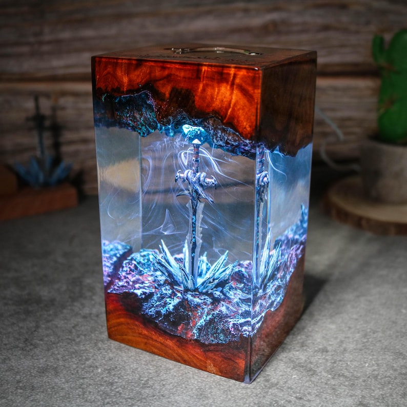 Puede incluir: Una caja de madera con un inserto de resina transparente que contiene una espada plateada. La resina est&aacute; llena de remolinos azules y morados. La caja est&aacute; iluminada desde el interior, creando un efecto brillante.