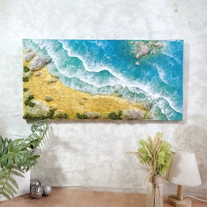 May include: A resin art painting depicting an aerial view of a beach with turquoise water and white foamy waves crashing on the shore. The beach is a light golden color with green foliage and small rocks.