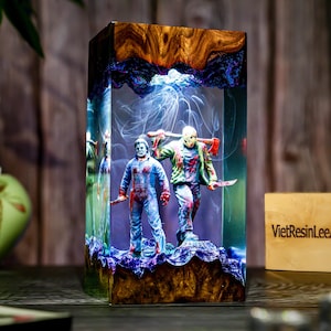 May include: A rectangular resin art piece featuring two horror movie figures with weapons. The figures are inside a clear resin block with a wood grain top and base. The piece has a blue and purple color scheme with the text "VietResinLeeArt".