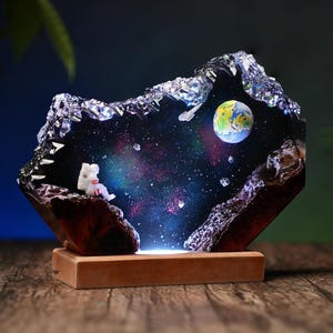 Könnte beinhalten: Dekorative Harz-Nachtlampe mit einem Miniatur-Astronauten, der eine farbenfrohe Galaxie mit einem Planeten betrachtet. Das Licht steht auf einem Holzsockel und schafft eine einzigartige und künstlerische Darstellung.
