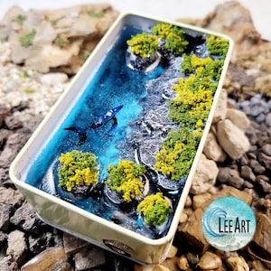 May include: A miniature ocean diorama in a rectangular container. The scene features a blue whale swimming in turquoise water, with miniature islands covered in yellow and green foliage. The container has a light green border.