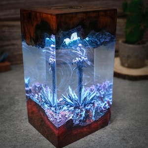 Puede incluir: Una base de madera con un bloque de resina transparente que contiene dos espadas y cristales azules y morados. La resina est&aacute; iluminada desde el interior, creando un efecto brillante.