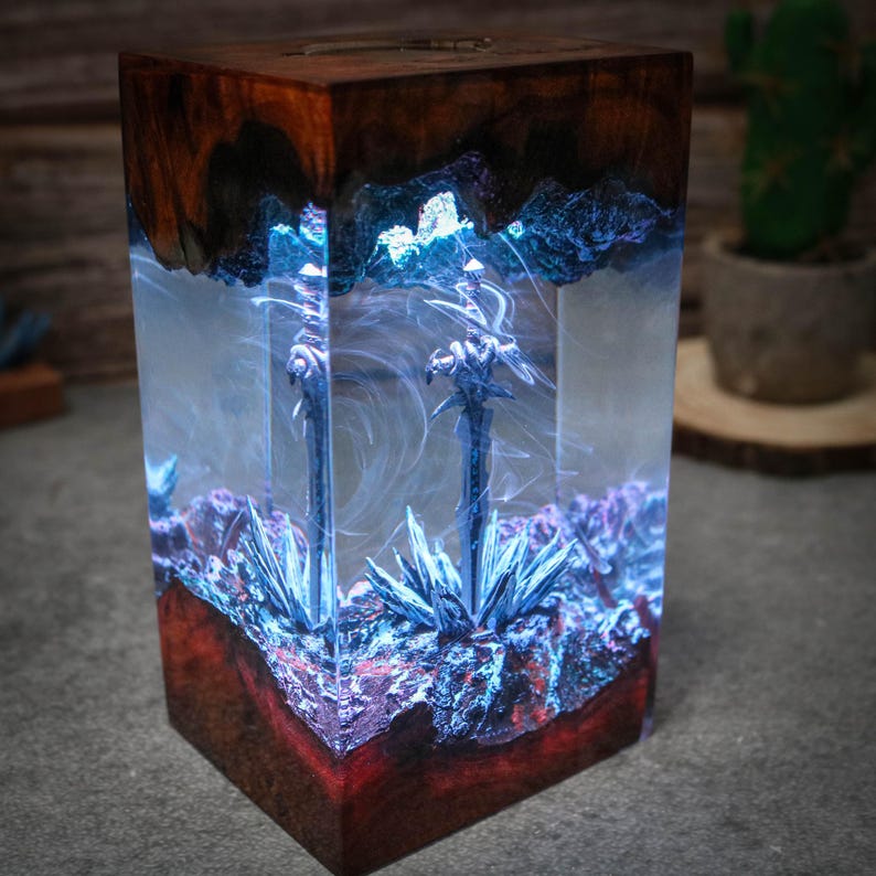 Puede incluir: Una base de madera con un bloque de resina transparente que contiene dos espadas y cristales azules y morados. La resina est&aacute; iluminada desde el interior, creando un efecto brillante.