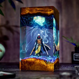 Puede incluir: Una pieza de arte de resina rectangular con una figura en miniatura con un casco con máscara roja y dos sables de luz. La figura se encuentra sobre una base texturizada dentro de un bloque de resina transparente con patrones arremolinados azules y blancos y un marco de madera.