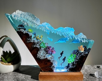 Könnte beinhalten: Eine Harzskulptur einer Meereswelle in Blau- und Grüntönen. Die Welle ist so geformt, dass sie aussieht, als würde sie über ein Korallenriff mit Fischen und Tauchern brechen. Die Skulptur ist auf einem Holzsockel montiert.