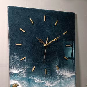 K&ouml;nnte beinhalten: Eine dunkelblaue Uhr mit goldenen Zeigern und goldenen Stundenmarkierungen. Das Zifferblatt der Uhr ist in ein harz&auml;hnliches Material eingebettet, das an Meereswellen erinnert.