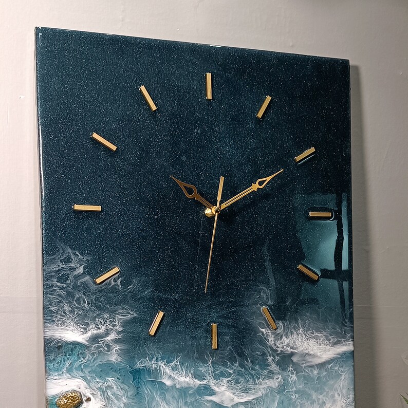 K&ouml;nnte beinhalten: Eine dunkelblaue Uhr mit goldenen Zeigern und goldenen Stundenmarkierungen. Das Zifferblatt der Uhr ist in ein harz&auml;hnliches Material eingebettet, das an Meereswellen erinnert.