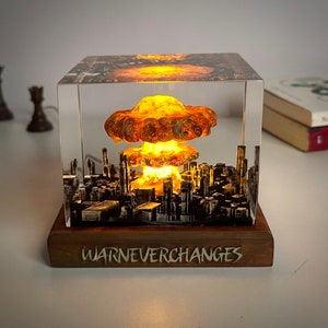 Könnte beinhalten: Ein transparenter Würfel mit einer Miniatur-Stadtlandschaft im Inneren, in der eine leuchtende orange-gelbe Pilzwolke explodiert. Der Würfel steht auf einem Holzsockel mit der Aufschrift "War Never Changes".