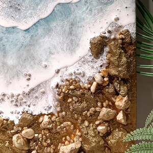 K&ouml;nnte beinhalten: Abstrakte Harz-Kunstmalerei, die eine Strandszene mit wei&szlig;en, schaumigen Wellen darstellt, die an einem goldenen Ufer mit kleinen wei&szlig;en und braunen Steinen brechen.