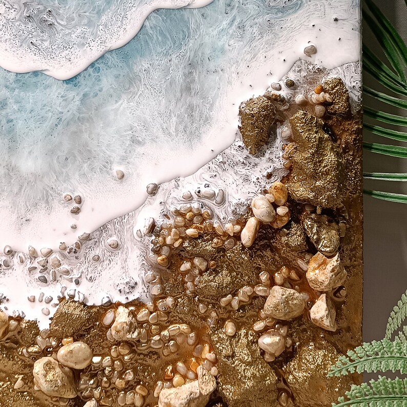 K&ouml;nnte beinhalten: Abstrakte Harz-Kunstmalerei, die eine Strandszene mit wei&szlig;en, schaumigen Wellen darstellt, die an einem goldenen Ufer mit kleinen wei&szlig;en und braunen Steinen brechen.