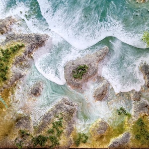 K&ouml;nnte beinhalten: Draufsicht auf eine K&uuml;stenlandschaft mit wei&szlig;en, sch&auml;umenden Wellen, die gegen Felsformationen schlagen. Das Wasser wechselt von T&uuml;rkis zu Wei&szlig;, mit gr&uuml;nen und braunen Akzenten. Kleine gr&uuml;ne Pflanzen bedecken die Felsen und bilden eine Miniaturlandschaft.