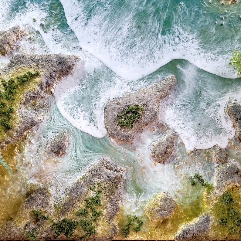 K&ouml;nnte beinhalten: Draufsicht auf eine K&uuml;stenlandschaft mit wei&szlig;en, sch&auml;umenden Wellen, die gegen Felsformationen schlagen. Das Wasser wechselt von T&uuml;rkis zu Wei&szlig;, mit gr&uuml;nen und braunen Akzenten. Kleine gr&uuml;ne Pflanzen bedecken die Felsen und bilden eine Miniaturlandschaft.